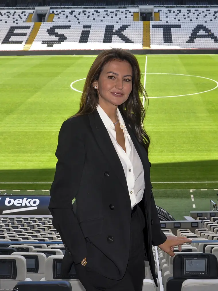 Mehtap Ferah, siyah takım elbisesiyle Beşiktaş Stadyumu’nun tribününde; arkasında saha ve koltuklarda “BEŞİKTAŞ” yazısı.