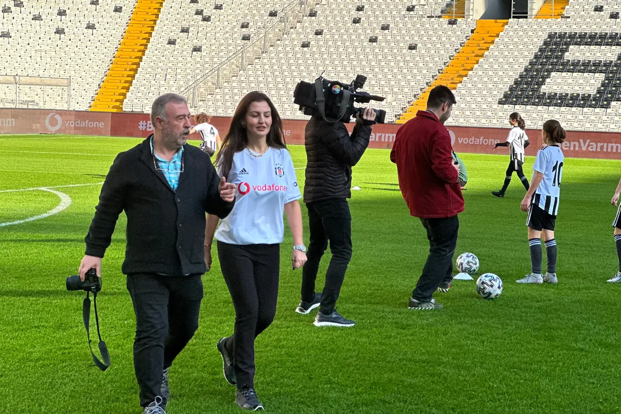 Mehtap Ferah, Beşiktaş Stadyumu’nda sahada yürüyor; yanında fotoğraf makinesi tutan bir kişi, arkasında kamera çeken bir ekip.