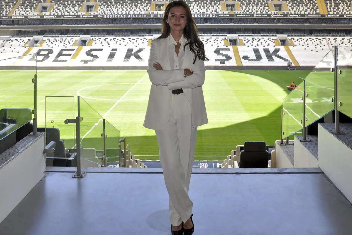 Mehtap Ferah, beyaz takım elbisesiyle Beşiktaş Stadyumu’nda tribün locasının önünde duruyor; arkasında saha ve koltuklarda “BEŞİKTAŞ JK” yazısı.